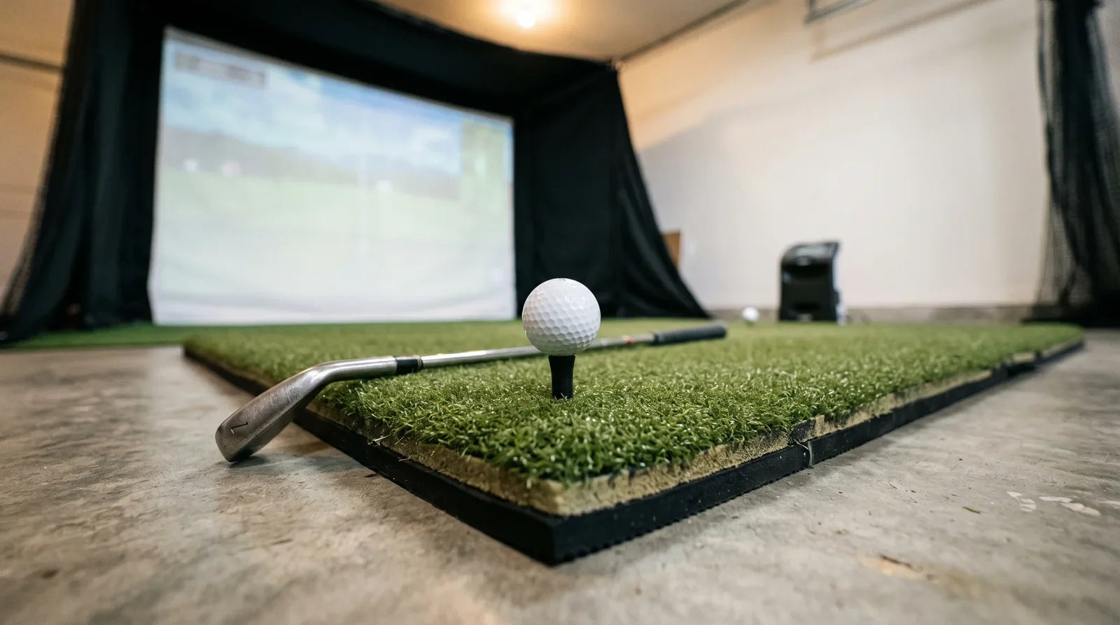 A close-up of a thick green golf hitting mat on a concrete garage floor in a home simulator, with a ball on a rubber tee and an iron resting on the turf